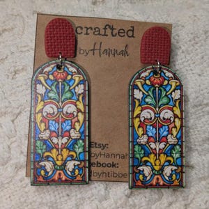 May include: A pair of colorful stained-glass style earrings with red tops. The earrings feature intricate floral and geometric designs in shades of blue, yellow, green, and red. The earrings are displayed on a brown card with the text "crafted by Hannah".