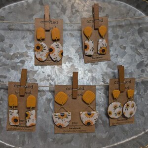 May include: A display of handmade earrings featuring a sunflower design. The earrings are attached to small brown cards with wooden clothespins. The earrings have a yellow and white color scheme, with a variety of shapes and sizes.