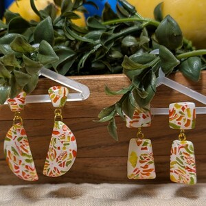 May include: Four pairs of colorful polymer clay earrings with gold-colored accents hang on clear plastic hangers. The earrings feature white, yellow, orange, and green patterns. The background includes lemons and greenery.