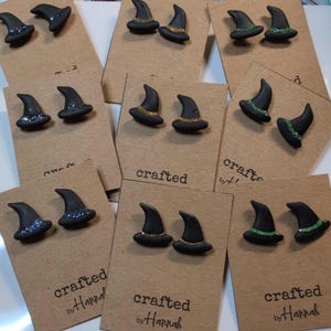 May include: Black witch hat earrings with glitter accents, displayed on brown cards. The earrings have a classic pointed shape and are decorated with silver, gold, or green glitter. The cards are labeled "crafted by Hannah".