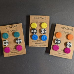 May include: Three sets of handcrafted stud earrings on brown cards. Each card displays six round earrings in various colors and patterns, including teal, yellow, black and white checkered, blue, orange, and pink. The cards have the text "crafted by Hannah".