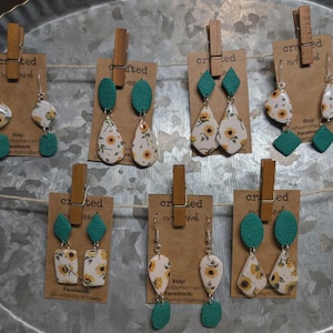 May include: A collection of handmade earrings with a sunflower pattern. The earrings feature a variety of shapes, including teardrops and squares, with teal accents. Each pair is displayed on a brown card with a wooden clip.