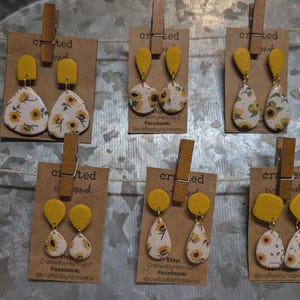 May include: A display of handmade earrings featuring a sunflower design. The earrings have yellow and white floral patterns, with yellow textured tops. Each pair is attached to a small brown card with a wooden clip. The earrings are displayed on a metallic surface.