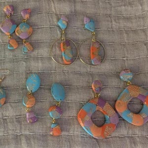 May include: A collection of colorful handmade earrings in various shapes and designs. The earrings feature a mix of blue, orange, purple, and gold tones. Styles include teardrop, geometric, and hoop earrings, all with unique patterns.