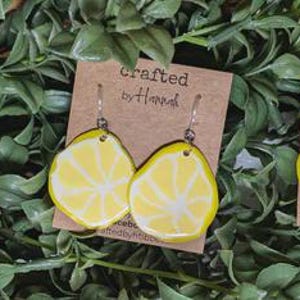 May include: Handmade lemon-shaped earrings with yellow and white details, displayed on brown tags with the text "Crafted by Hand." The earrings are set against a backdrop of green foliage, showcasing a fresh, summery aesthetic.