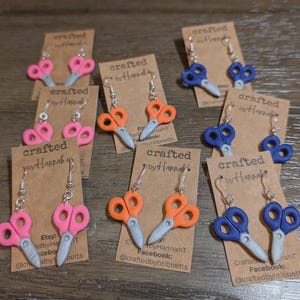 May include: A collection of handmade scissor earrings in various colors, including pink, orange, and blue. Each pair is attached to a small, brown card with the text "crafted by Hannah" and social media information. The scissors have silver-colored blades.