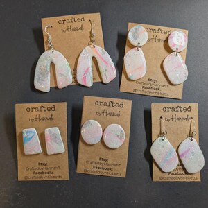 May include: A collection of handmade polymer clay earrings in various shapes and designs. The earrings feature a marbled effect with pastel pink, blue, and silver accents. Each pair is displayed on a brown card with the text "crafted by Hannah".