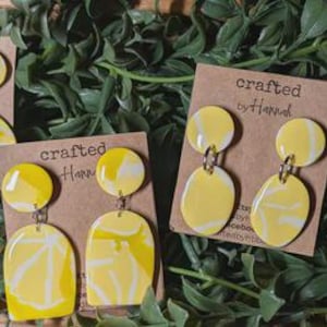May include: A collection of handmade yellow and white marbled earrings on brown tags. The earrings feature various shapes, including arches, circles, and ovals. The tags read "crafted by hand."