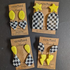 May include: Four pairs of handmade earrings on display. Each pair features a yellow accent, with designs including hearts, stars, and lightning bolts. The earrings have a black and white checkered pattern. The cards read "crafted by Hannah".