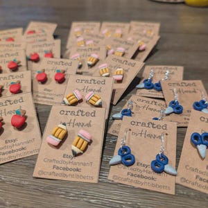 May include: A collection of handmade earrings on display. The earrings feature designs of red apples, yellow pencils, and blue scissors. Each pair is mounted on a small, brown card with the text "crafted by Hannah" and social media information.