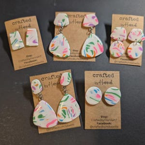 May include: Five pairs of handmade polymer clay earrings. Each pair features a white base with colorful abstract designs in pink, green, blue, and orange. The earrings are displayed on small brown cards with the text "crafted by Hannah".