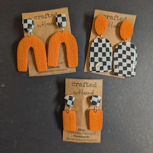 May include: Three pairs of handmade earrings. One pair features orange arches, another has black and white checkered shapes, and the third has orange rectangles. Each pair has a black and white checkered top. The earrings are displayed on brown cards with the text "crafted by Hannah".