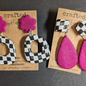 May include: Two pairs of handmade earrings. One pair features a flower-shaped magenta top and a black and white checkered oval. The other pair has a checkered square top and a magenta teardrop shape. The earrings are on brown cards.