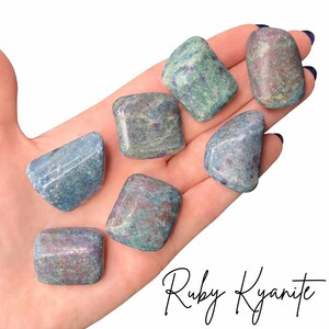 May include: Seven polished ruby kyanite stones in varying shades of blue, green, and red are displayed in a hand. The stones have a smooth, rounded shape. The text "Ruby Kyanite" is written in cursive script at the bottom of the image.