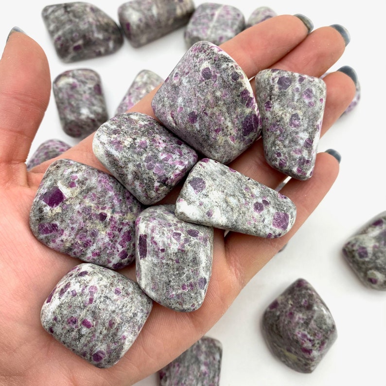 May include: A collection of tumbled ruby in kyanite stones. The stones are a mix of grey and pink with a speckled appearance.