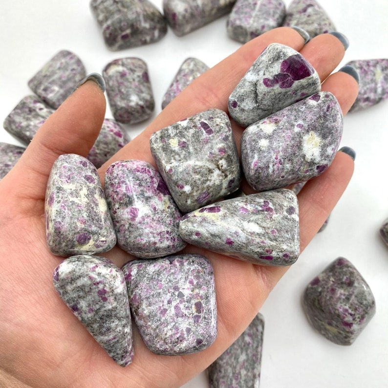 May include: A collection of tumbled ruby in kyanite stones. The stones are a mix of grey and white with red ruby inclusions. The stones are smooth and polished.
