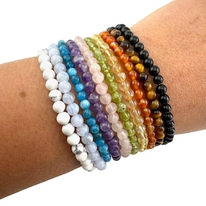 May include: A stack of 10 colorful beaded bracelets on a person's wrist. The bracelets are made of various gemstones, including white, blue, green, purple, pink, yellow, orange, and black.