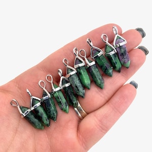 May include: A group of 12 green and black gemstone pendants with silver settings. The pendants are in the shape of a point and are arranged in a row on a hand.