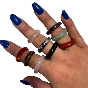 Puede incluir: Una mano exhibe una colección de anillos de piedras preciosas en varios colores. Los anillos incluyen piedras rojas, negras, verdes, moradas y transparentes. Los anillos se usan en varios dedos, mostrando sus diversos tonos y texturas.