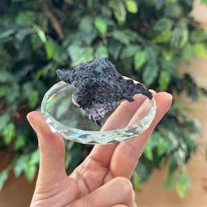 May include: A rough, dark charcoal-colored mineral specimen with a porous, jagged texture and subtle purple crystalline inclusions, resting on a clear, circular, faceted glass display base that reflects light along its beveled edges.
