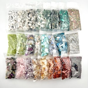 Op de afbeelding: Vierentwintig kleine plastic zakjes gevuld met verschillende soorten gekleurde edelsteenchips. De zakjes zijn gerangschikt in vier rijen van zes zakjes.