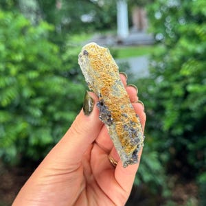 May include: A long, slender mineral specimen with a rough, textured surface. The stone features a prominent band of vibrant orange-yellow crusting along its centre, contrasted by dark, crystalline grey and black mineral deposits on the edges.