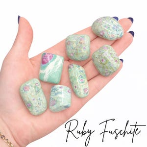 May include: Seven polished ruby fuchsite stones in varying shapes and sizes are displayed in a hand. The stones are primarily green with pink and red inclusions. The text "Ruby Fuschite" is written in a script font at the bottom of the image.