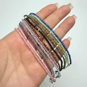 May include: A collection of delicate beaded bracelets in various colors, including pink, purple, blue, and black. Each bracelet features small, round beads strung together with a silver clasp. The bracelets are displayed together, showcasing their diverse hues.