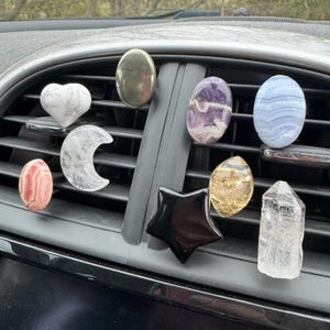 May include: A collection of decorative car air vent diffusers in various shapes and colors. Includes a white heart, a silver oval, a purple oval, a blue oval, a pink oval, a clear crescent moon, a black star, a yellow oval, and a clear crystal.