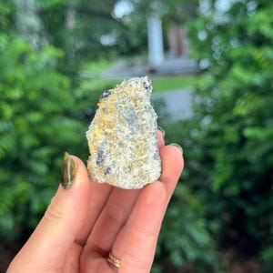 May include: A raw mineral specimen featuring a cluster of small, translucent, needle-like crystals embedded in a textured, light grey and golden-yellow matrix. The stone has an irregular, jagged shape with visible crystalline formations throughout.