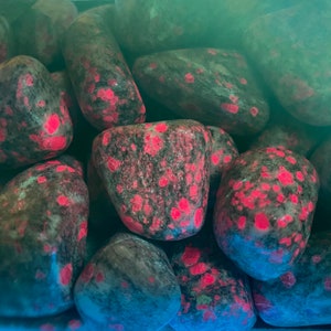 May include: Close-up of a group of tumbled stones with a dark grey base and bright pink flecks. The stones are smooth and polished.