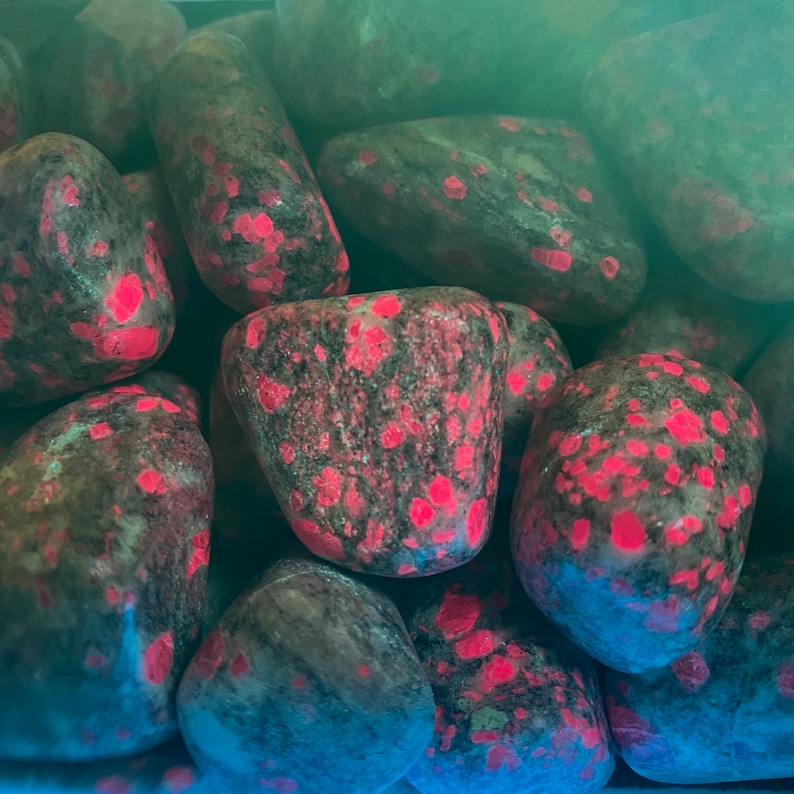 May include: Close-up of a group of tumbled stones with a dark grey base and bright pink flecks. The stones are smooth and polished.