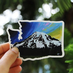 May include: A sticker of the state of Washington with a silhouette of Mount Rainier and a colorful sky in the background.