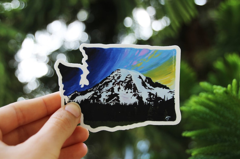 May include: A sticker of the state of Washington with a silhouette of Mount Rainier and a colorful sky in the background.