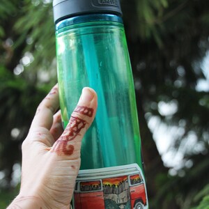 May include: A green water bottle with a black lid and a sticker of a red vintage van with a mountain scene. The bottle is being held by a hand with henna.