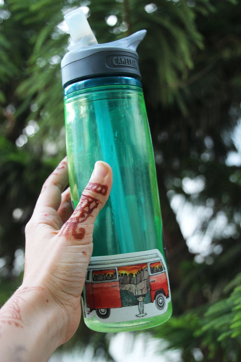 May include: A green water bottle with a black lid and a sticker of a red vintage van with a mountain scene. The bottle is being held by a hand with henna.