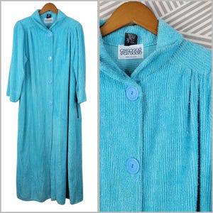 May include: A bright turquoise, long-sleeved, floor-length robe made of a textured, ribbed fabric. It features a collared neckline, four matching turquoise buttons down the front, and a label at the neck that reads TUFTEES MADE IN U.S.A.