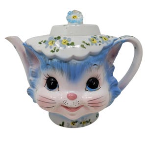 May include: A ceramic teapot shaped like a cat head with blue fur, large black eyes, a pink nose, and whiskers. The lid features a small blue flower handle. The body is decorated with painted blue flowers and green leaves on a white background.