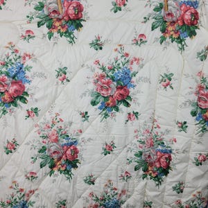 May include: Cream-colored floral quilt featuring a repeating pattern of large pink and red roses, blue hydrangeas, and green leaves. The quilt has a quilted design with a diamond pattern.