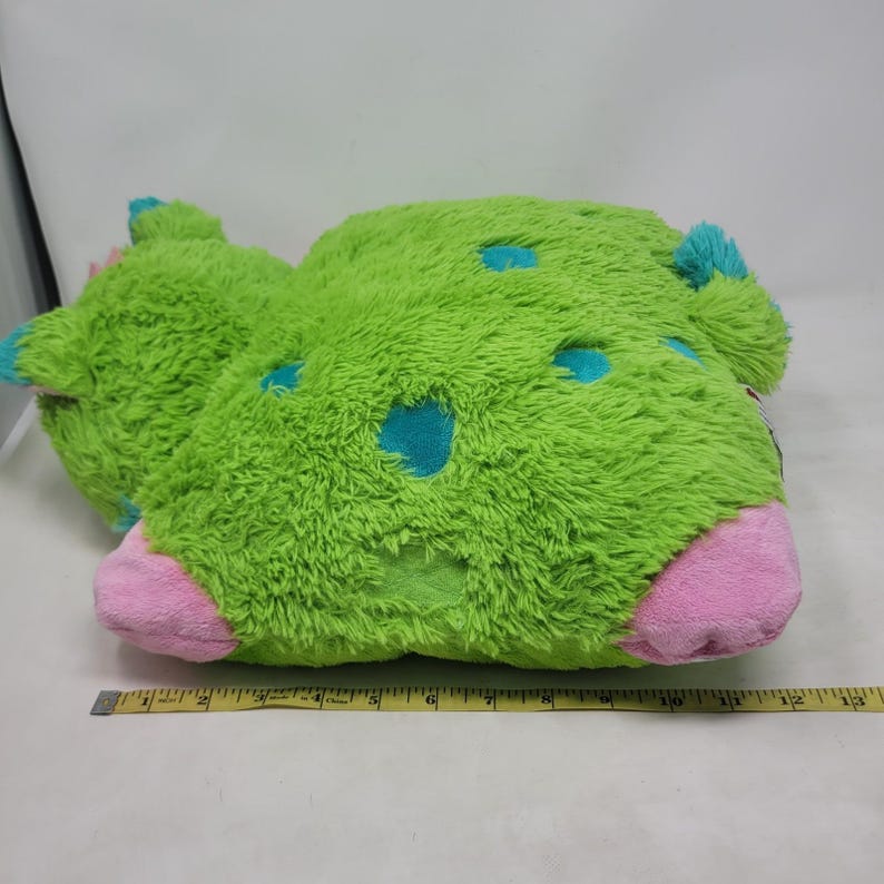 Puede incluir: Una almohada de felpa verde con forma de cactus con detalles azules y rosas. La almohada tiene una textura peluda y manchas azules. La base es rosa y las puntas son azules. Una cinta m&eacute;trica est&aacute; colocada junto a la almohada.