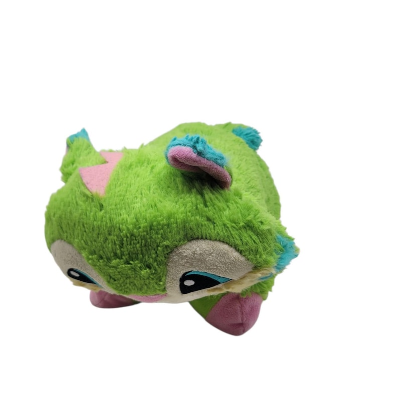 Puede incluir: Un peluche verde con forma de criatura, con detalles rosas y azules. El juguete tiene ojos grandes y una textura suave y peluda. Se ven tri&aacute;ngulos rosas y detalles azules en las orejas y el cuerpo.