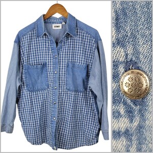 May include: A denim shirt with a blue and white check pattern on the front and denim sleeves. The shirt features two front pockets and a collar. The sleeves are a lighter shade of blue denim. The shirt is displayed on a wooden hanger.