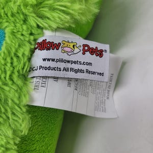 Puede incluir: Primer plano de un juguete de peluche verde con una etiqueta. La etiqueta dice "Pillow Pets" con un logotipo de vaca y el sitio web www.pillowpets.com. La etiqueta tambi&eacute;n incluye informaci&oacute;n sobre los materiales y el origen del producto.