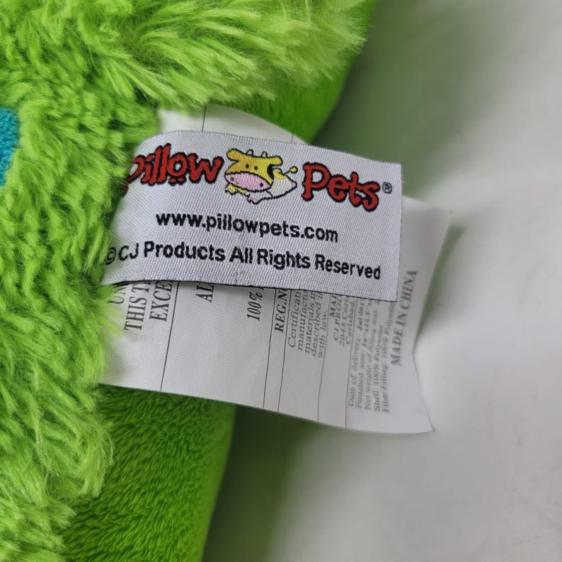 Puede incluir: Primer plano de un juguete de peluche verde con una etiqueta. La etiqueta dice "Pillow Pets" con un logotipo de vaca y el sitio web www.pillowpets.com. La etiqueta tambi&eacute;n incluye informaci&oacute;n sobre los materiales y el origen del producto.
