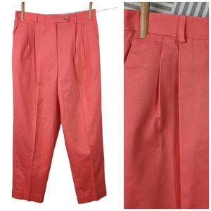 May include: A pair of coral-colored pleated pants with a button and zipper closure. The pants have a straight leg and a cuffed hem.