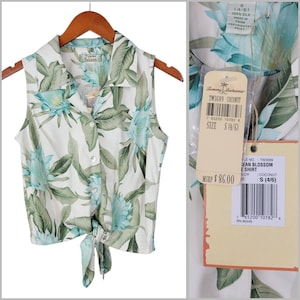 May include: A sleeveless white button-down shirt featuring a green and light blue tropical floral print. The shirt has a collared neckline and a tie-front hem. A tag attached to the garment displays the text Tommy Bahama and size S (4/6).