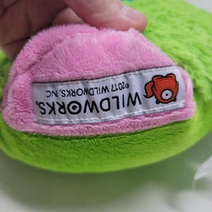 Puede incluir: Primer plano de un juguete de peluche con cuerpo verde y parte inferior rosa. Una etiqueta blanca con el texto "WILDWORKS" y un personaje de dibujos animados est&aacute; cosida a la tela rosa. La etiqueta tambi&eacute;n incluye el texto "&copy;2017 WILDWORKS, INC."