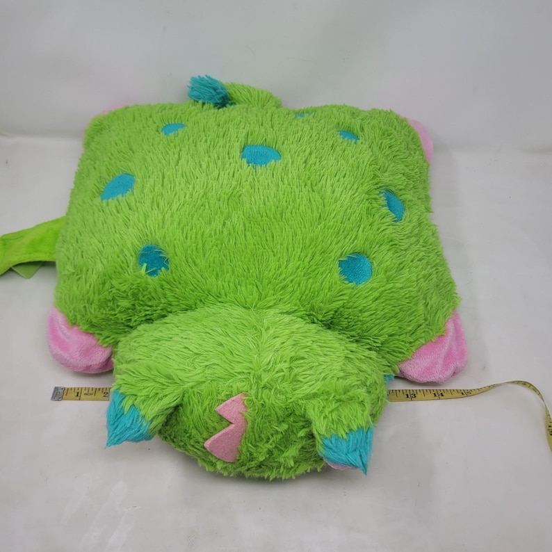 Puede incluir: Una almohada de felpa verde, de forma cuadrada, con lunares azules, detalles rosas y garras azules. La almohada tiene una textura suave y peluda y un dise&ntilde;o caprichoso, que se asemeja a un monstruo o criatura amigable.