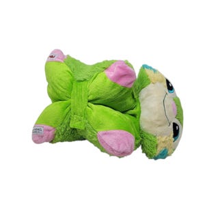 Puede incluir: Un juguete de peluche verde con detalles rosas en los pies y una cara de color claro. El juguete tiene ojos negros y detalles azules y amarillos. El juguete est&aacute; tumbado boca arriba.