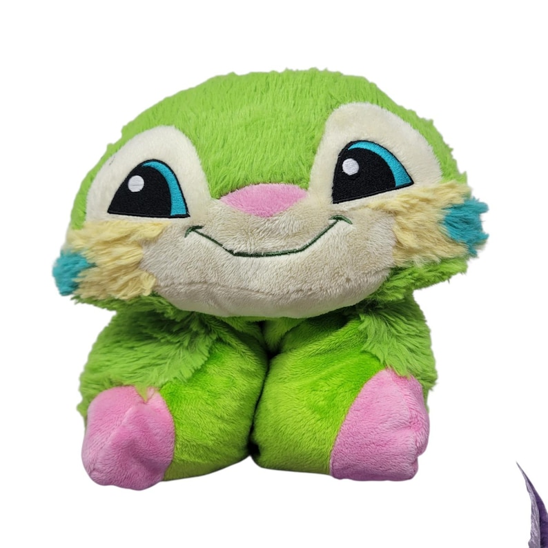 Puede incluir: Un peluche verde con forma de criatura sonriente. Tiene grandes ojos negros con detalles azules, una nariz rosa y patas rosas. Las orejas tienen detalles amarillos y azules. Juguete de peluche.